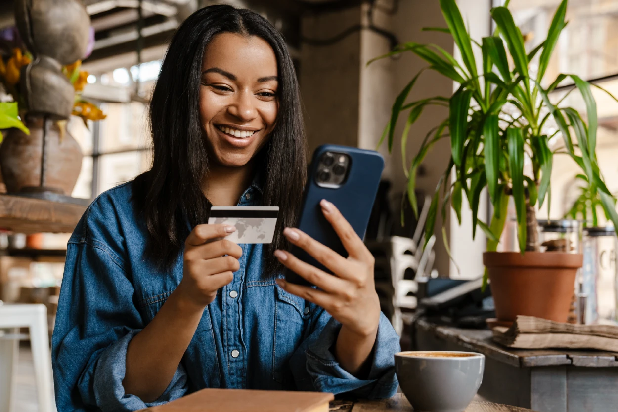 Mujer sonriente utilizando su teléfono móvil mientras sostiene una tarjeta bancaria en una mesa con una taza de café