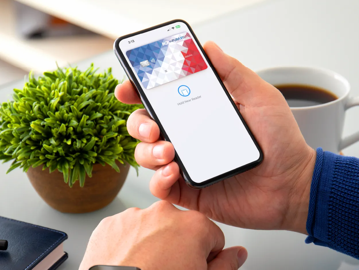 Persona sosteniendo un teléfono móvil que muestra una tarjeta digital del BCR en pantalla, sobre una mesa con una planta y una taza de café.