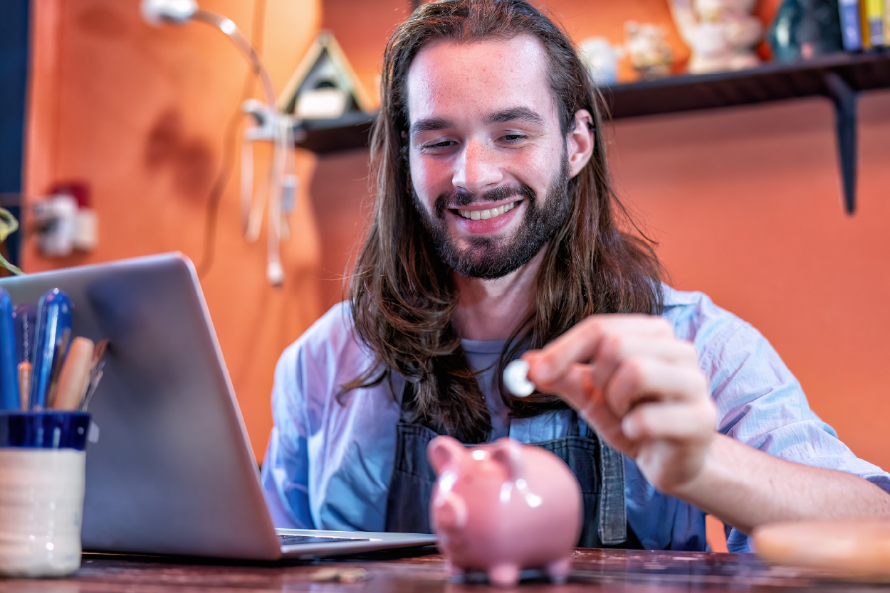“Hombre sonriente depositando una moneda en una alcancía rosada mientras usa una laptop en un escritorio.