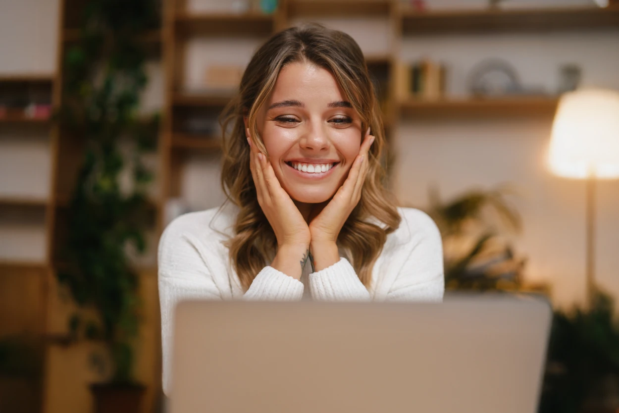 Mujer sonriente sentada frente a una computadora portátil, apoyando el rostro con las manos en un ambiente interior acogedor con plantas y una lámpara encendida.