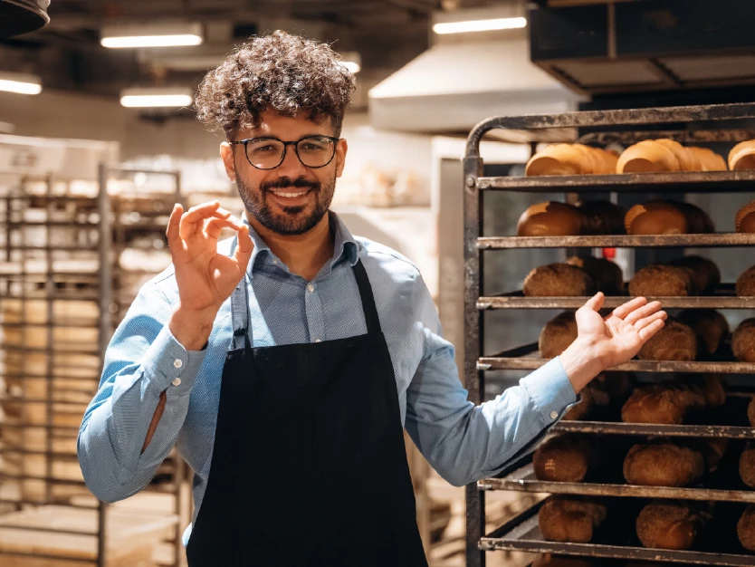 Marcos, dueño de una panadería, mostrando satisfacción junto a los estantes de pan en su negocio.